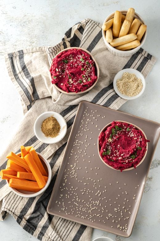 Overhead shot of beetroot hummus served in small white bowls with some crisp breads, carrot sticks and sesame seeds in the background