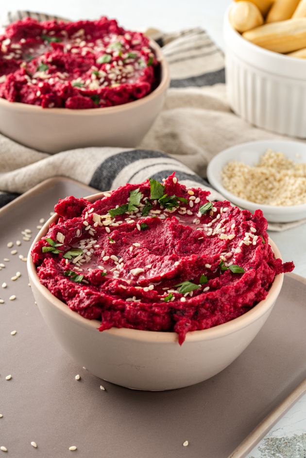 A bright pink dip served in a white bowl with a smaller bowl of sesame seeds and some crisp breads in the background