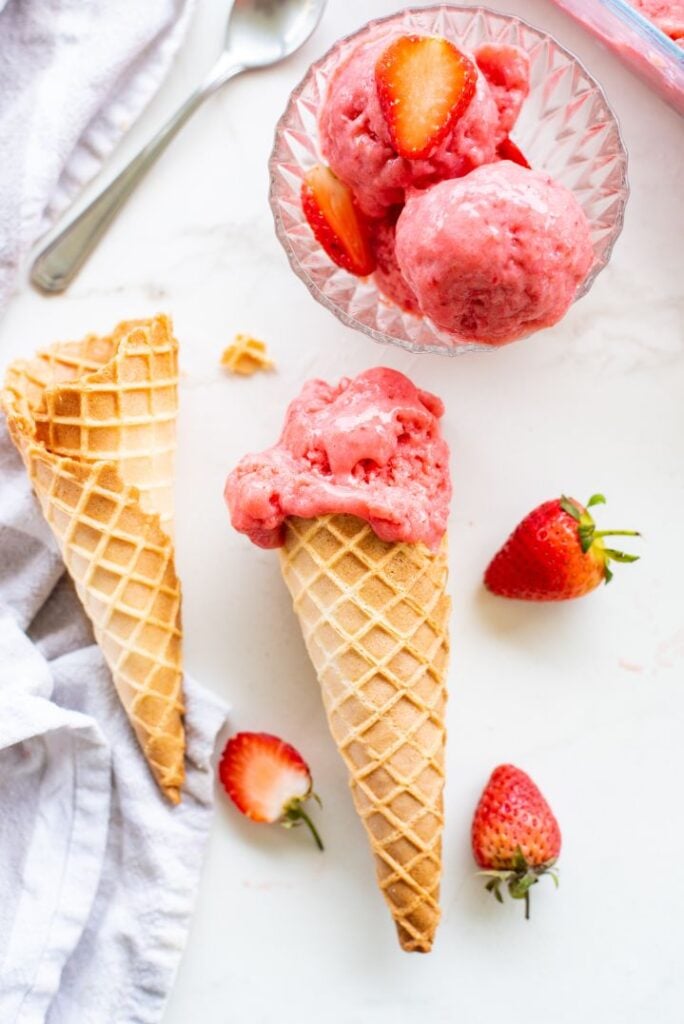 Single scoop of strawberry sorbet in a waffle cone with a bowl of sorbet beside it and some fresh strawberries