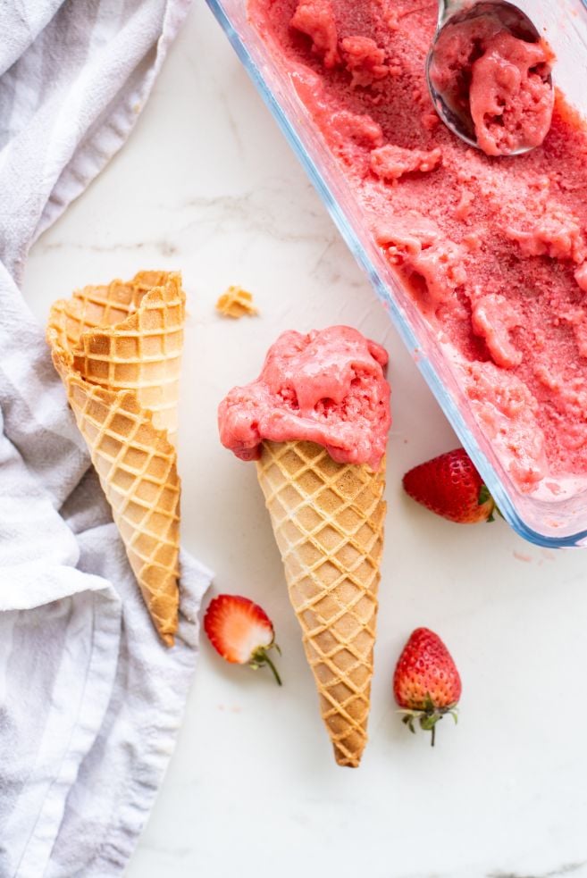 Overhead shot of strawberry sherbet in a glass container with a scoop of ice cream in an ice cream scoop, there are 2 waffle cones and some fresh strawberries on the side