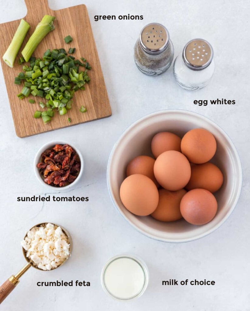 Overhead ingredient shot containing eggs, green onions, sundried tomatoes, feta cheese, milk, salt and pepper