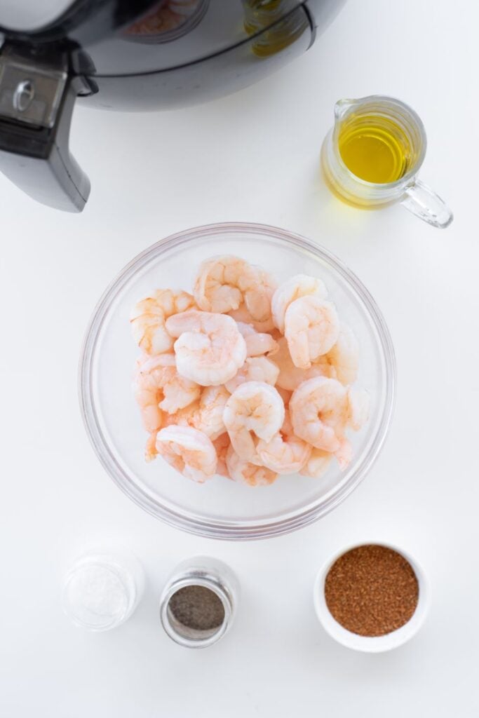 Overhead shot of pink shrimp in a glass bowl with cajun seasoning, olive oil, salt and pepper in small containers on the side.