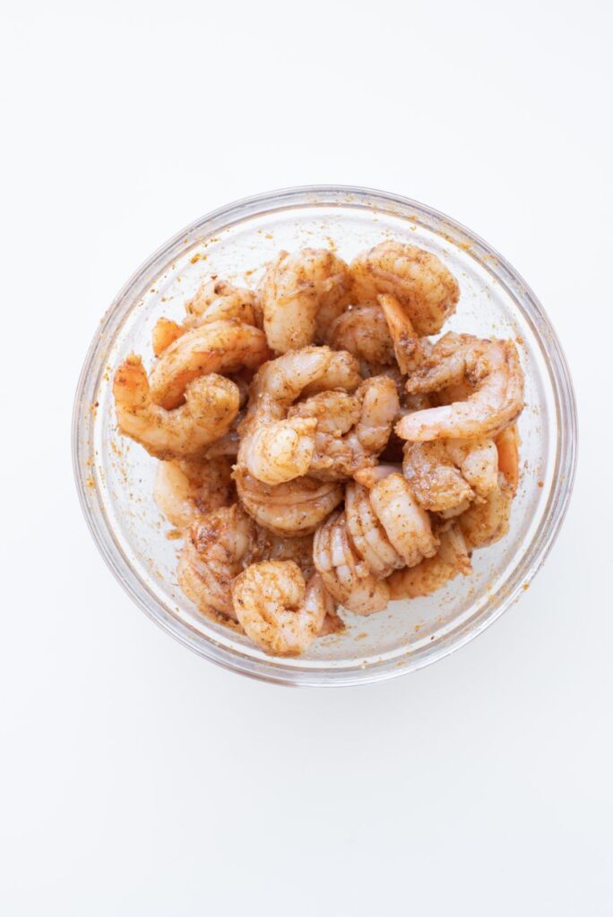 Overhead shot of uncooked shrimp in a glass bowl and covered in oil and seasoning.
