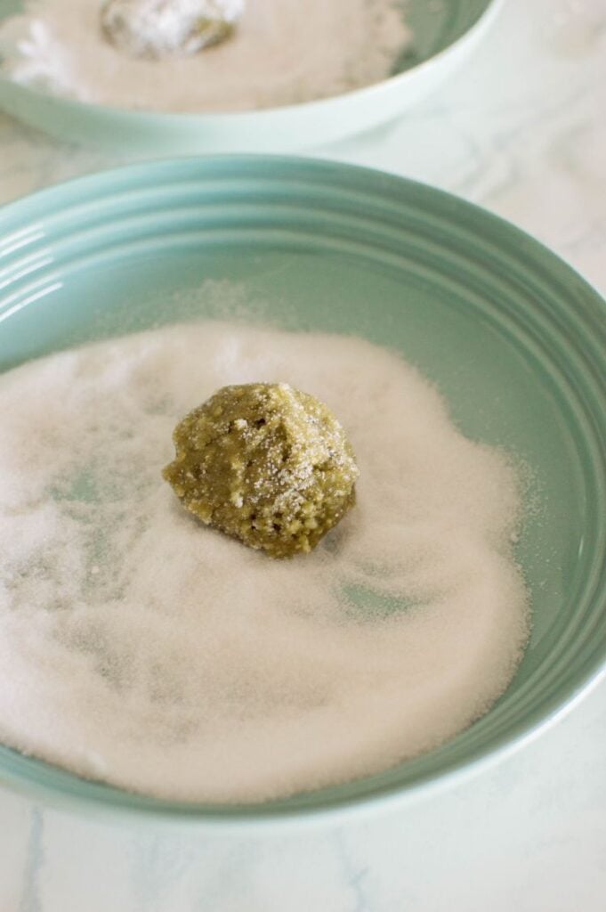 A small ball of green cookie dough being rolled in granulated sugar