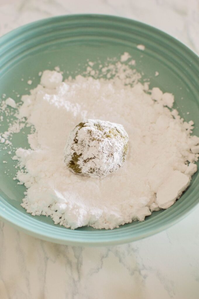 A small ball of green cookie dough being rolled in powdered sugar