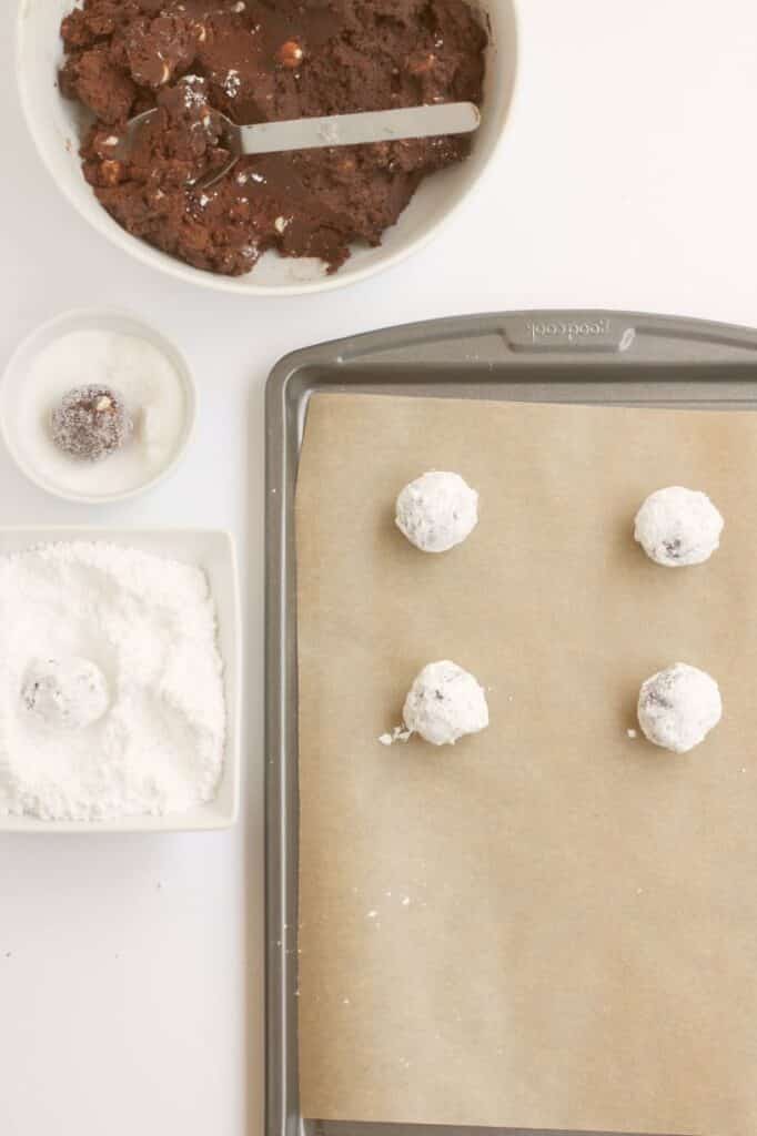 crinkle cookies being rolled into balls, dipped into granulated sugar and then powdered sugar and placed on a baking sheet