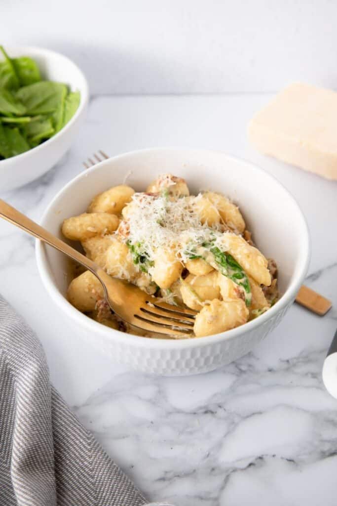 Front shot of a creamy pesto gnocchi recipe dish served up in a white bowl with a gold fork scooping it out