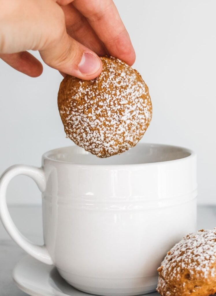 an orange colored cookie topped with powdered sugar being dipped into a mug containing tea