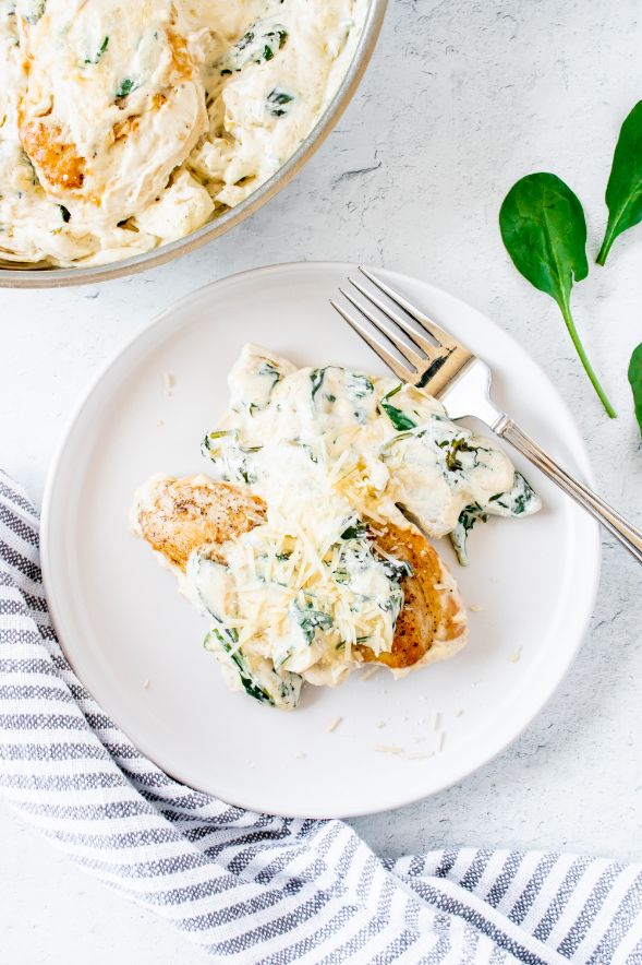 Creamy chicken breasts being served from a skillet to a small white plate and being sprinkled with freshly grated parmesan cheese