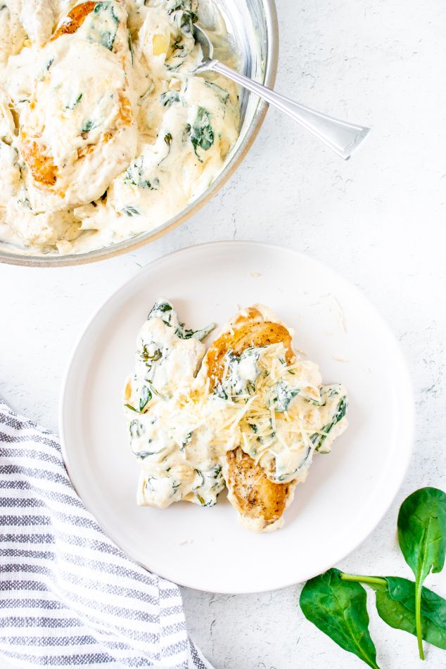 Creamy chicken breasts being served from a skillet onto a white plate with some baby spinach sprinkled in the background