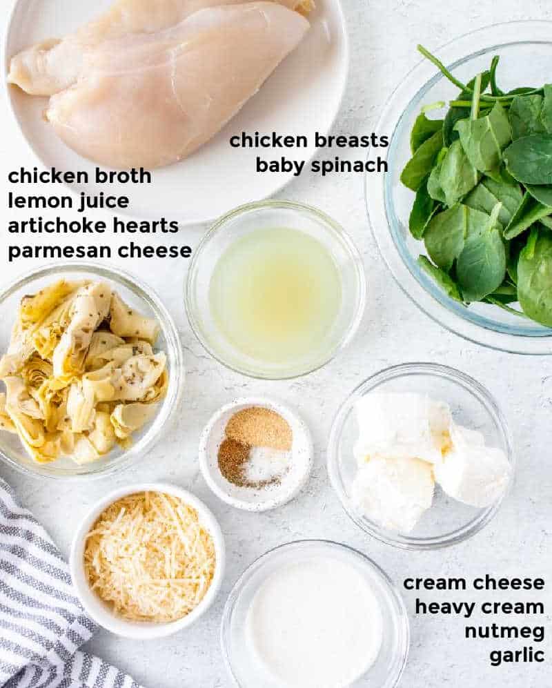 Overhead ingredient shot of chicken breasts, spinach, artichokes, parmesan cheese, heavy cream, cream cheese and spices served in small bowls