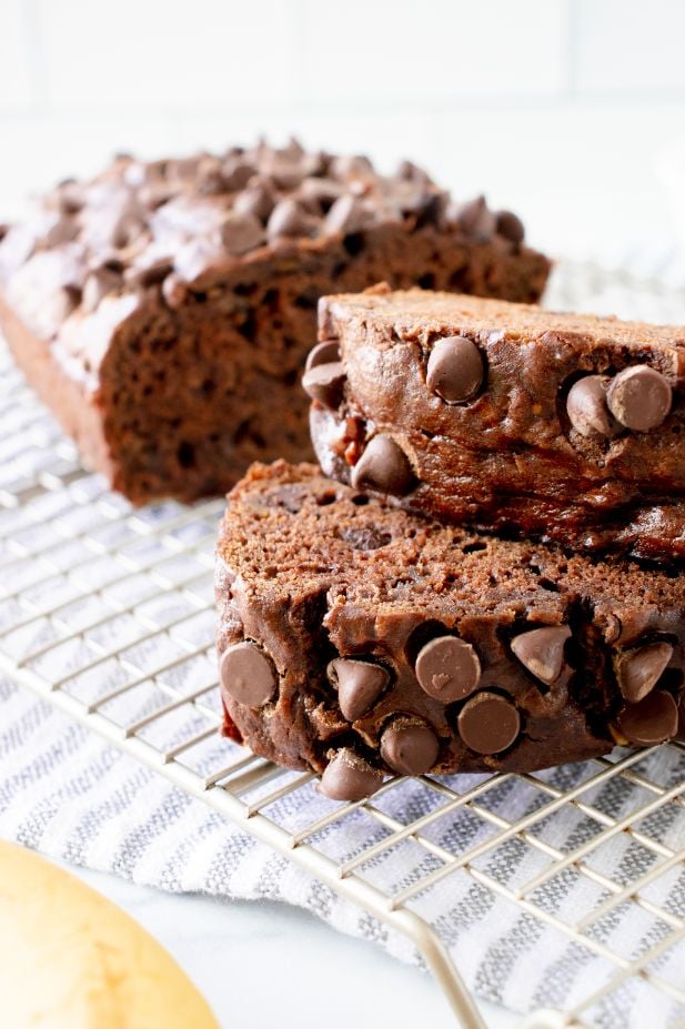 A stack of two slices of double chocolate banana bread cooling on a wire rack with the rest of the loaf in the background