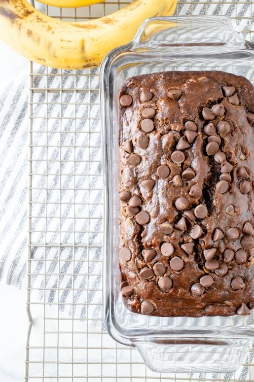 A baked banana bread cooling in a glass loaf pan on top of a wire rack with a banana in the background