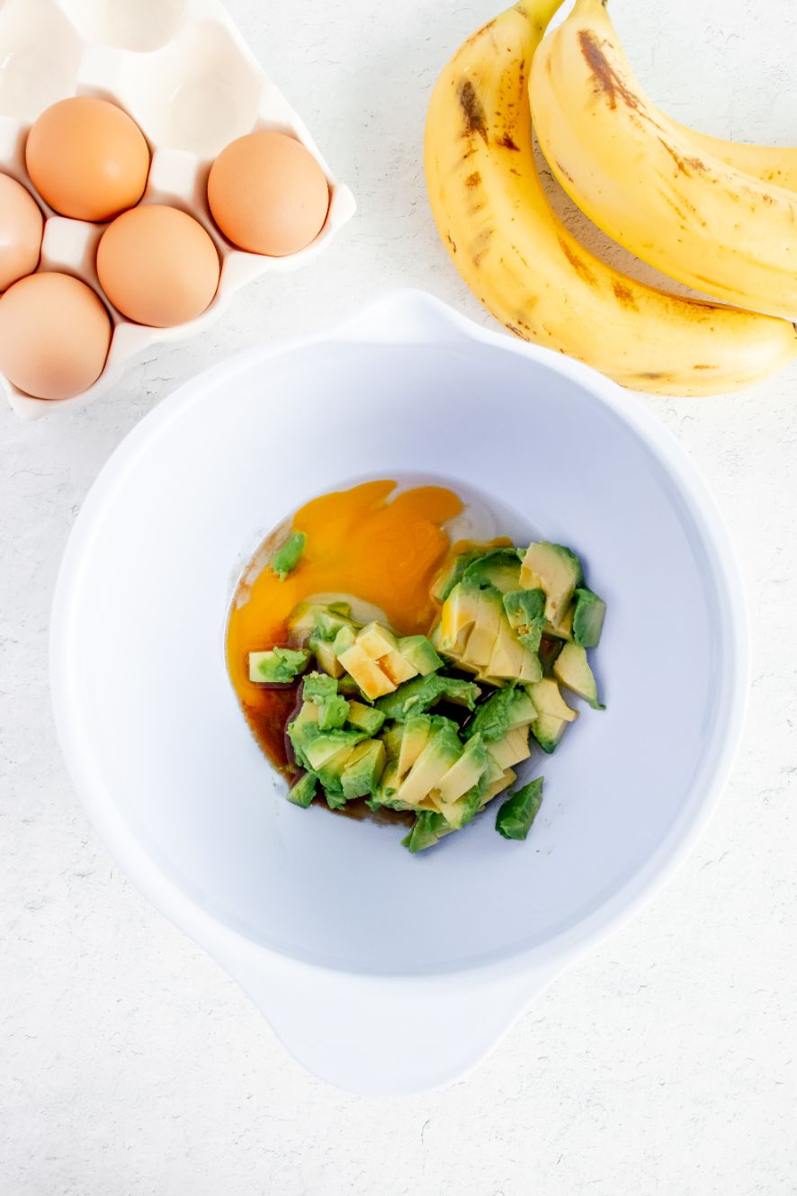 Overhead shot of avocado egg and vanilla in a white bowl with bananas in the background