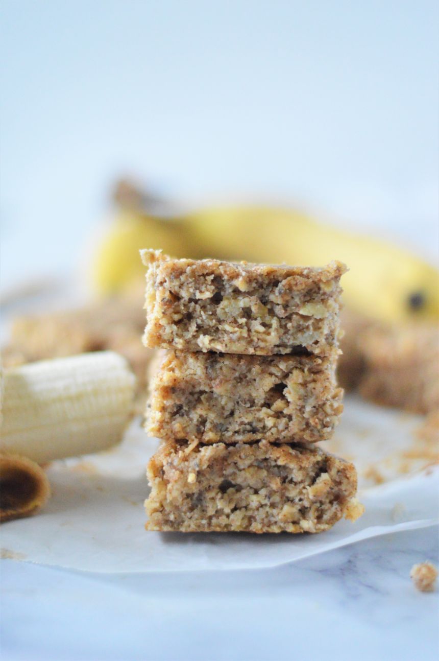 A stack of banana bread bars on some parchment paper with ripe bananas in the background
