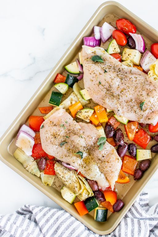 Seasoned chicken layered on top of Greek vegetables on a baking sheet