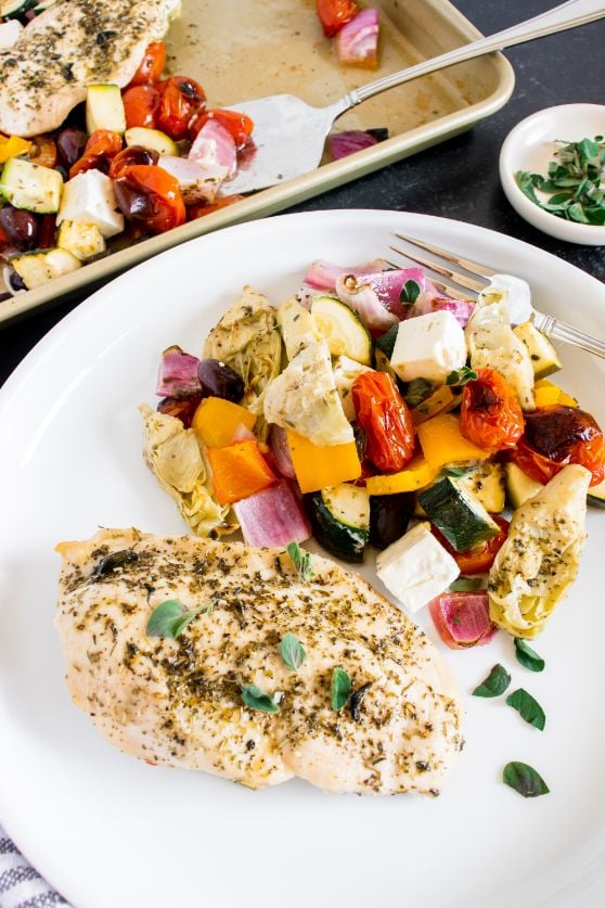 Greek chicken and vegetables served from the baking sheet to a white plate with a small bowl of fresh oregano in the background