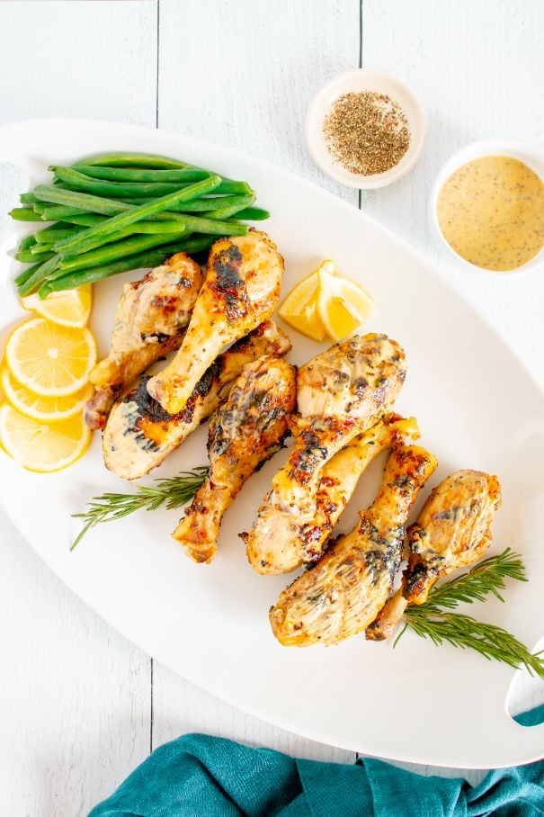 Overhead shot of grilled chicken drumsticks arranged on a white platter with a side of green beans, lemon slices and extra mustard
