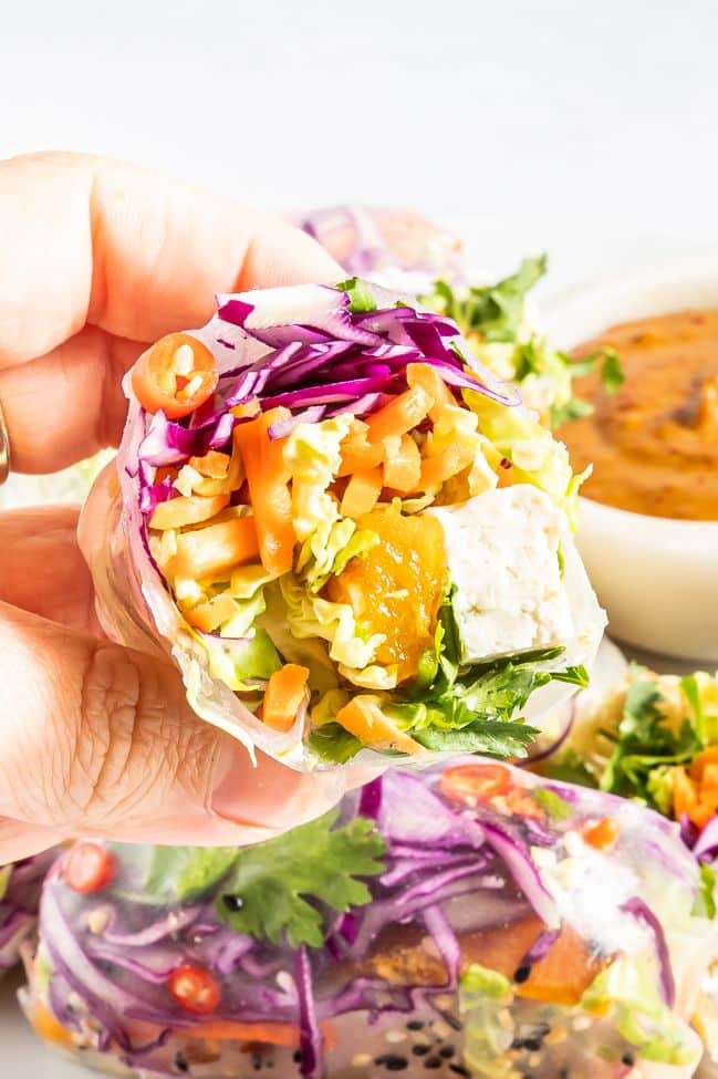 Hand holding a colorful vegetarian summer roll with pumpkin and tofu and there is a small container of dipping sauce in the background