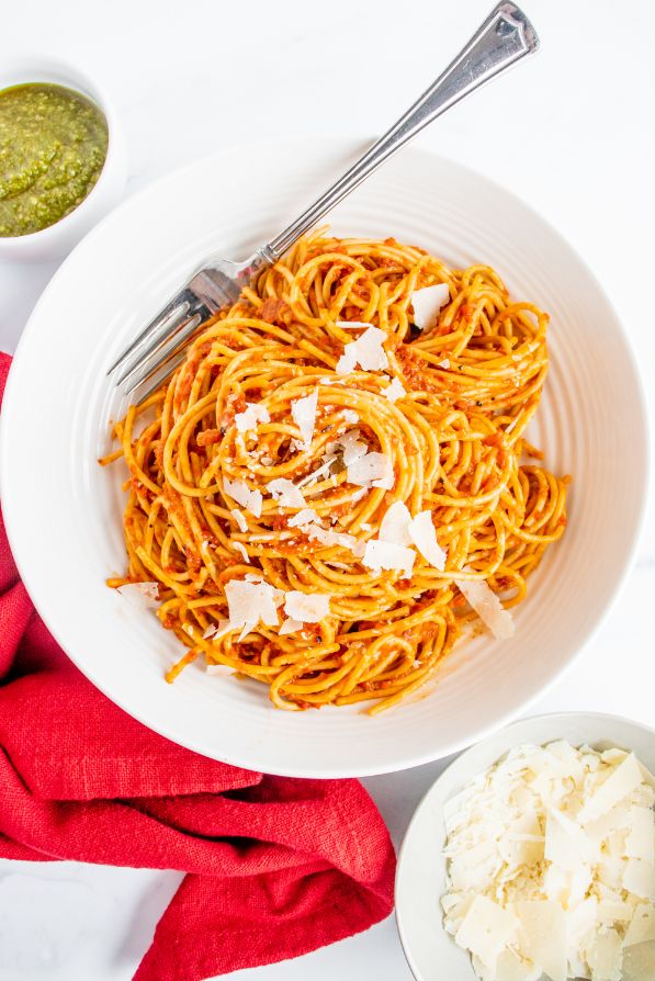 Red pepper pasta served in a white bowl with a small bowl of parmesan and another small bowl of pesto on the side