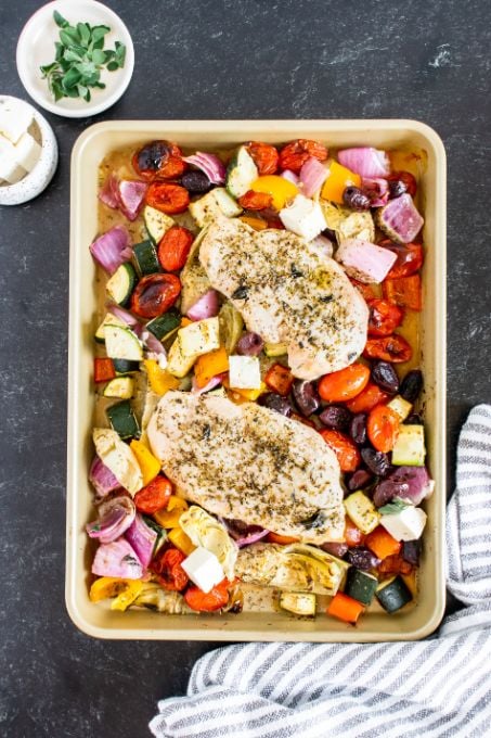 Cooked greek chicken and vegetables resting on a baking tray while fresh feta is sprinkled on