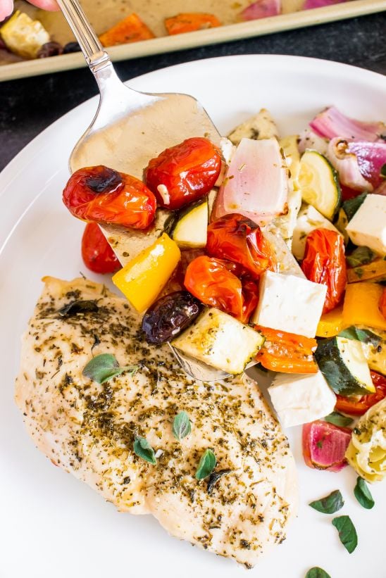 Sheet pan Greek chicken and veggies being served from the baking tray onto a white plate with sprinkles of fresh chopped oregano