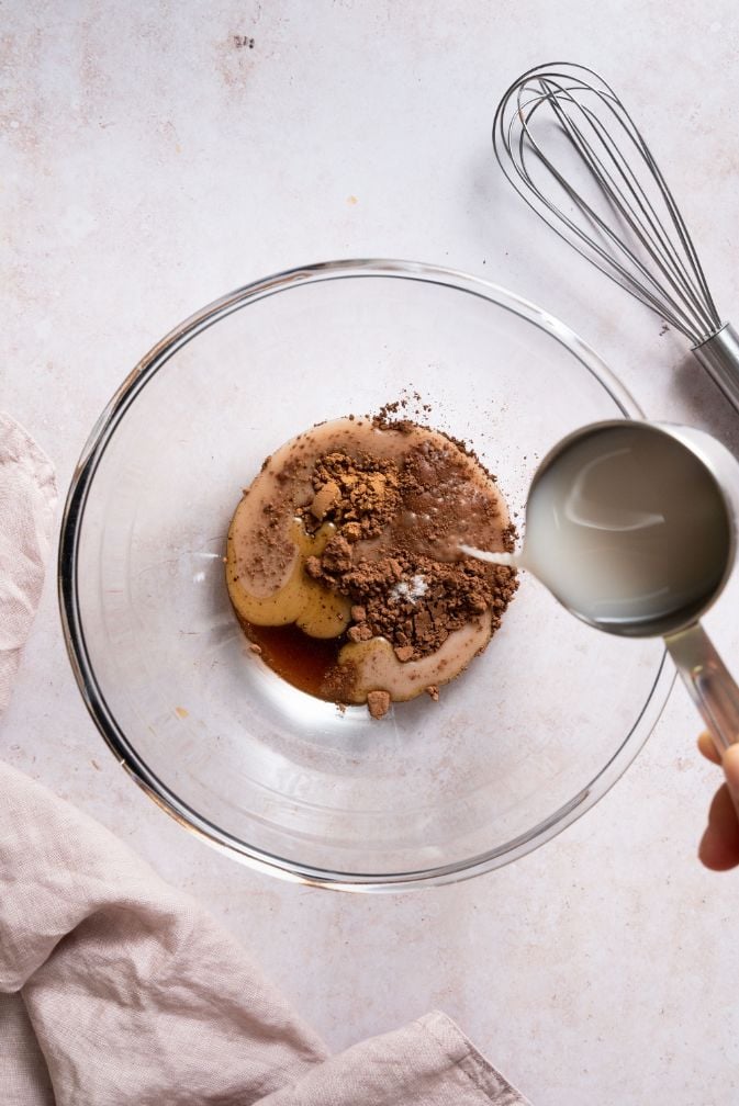 Large glass bowl containing cocoa, maple syrup, vanilla and sea salt with milk being poured in from above