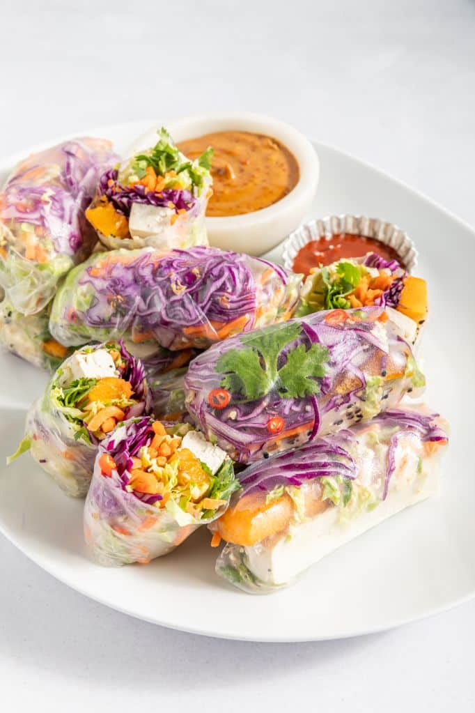 A bunch of colorful rice paper rolls arranged on a white platter plate with 2 dipping sauces on the side.