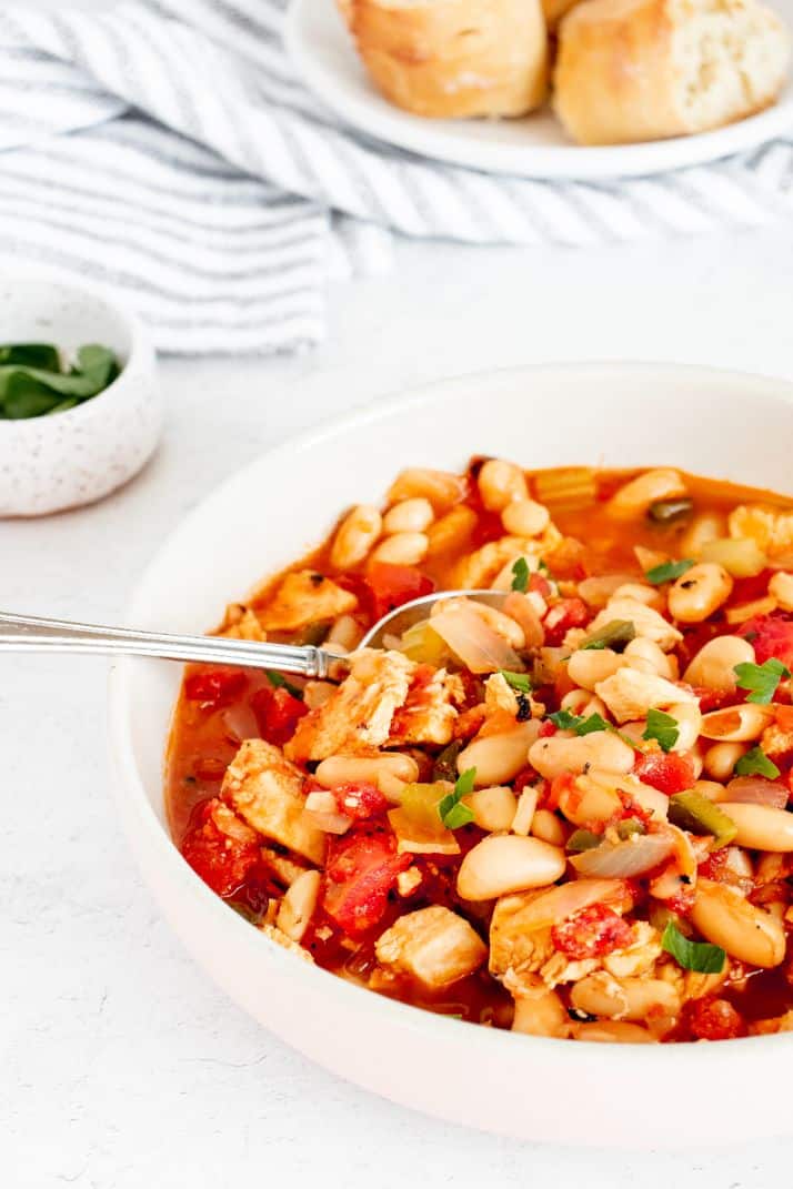 a white bowl with a spicy chicken and butter bean stew being spooned out. There is some fresh crusty bread and parsley in the background