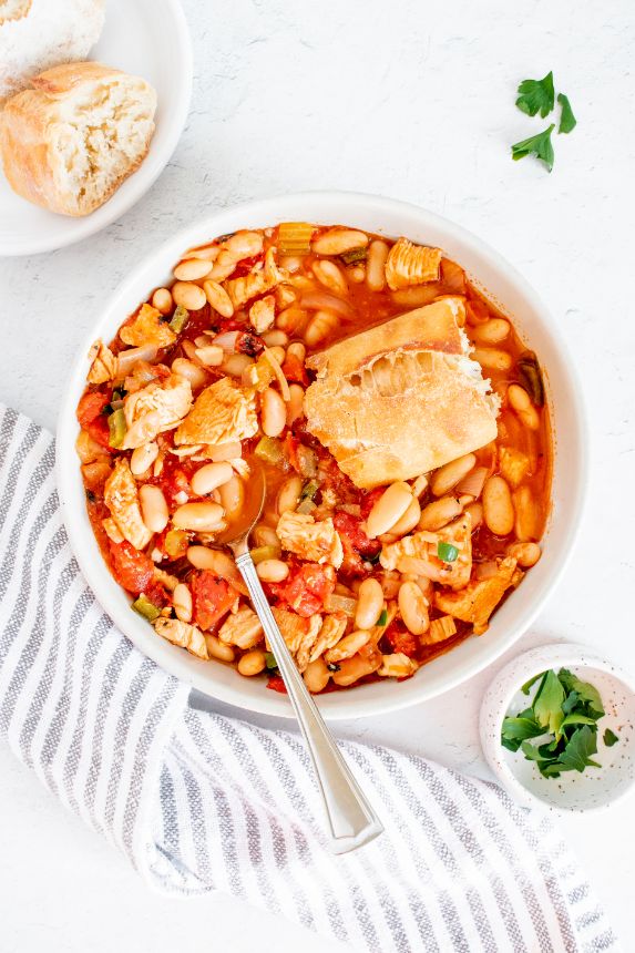 A white bowl containing a red tomato based stew and topped with some crusty bread and a silver spoon