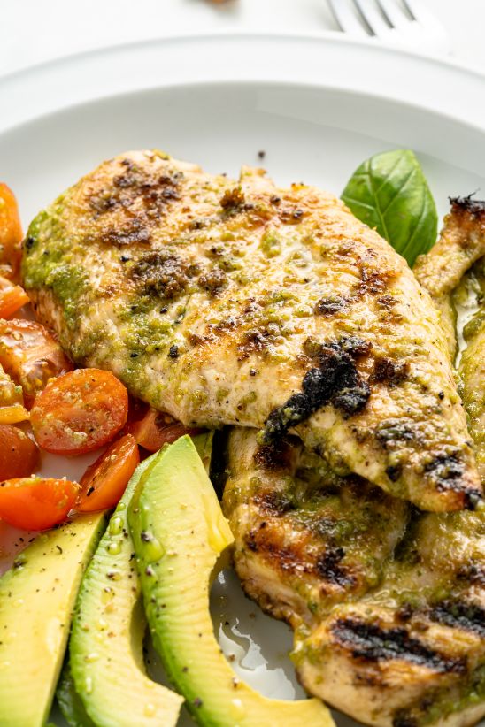 Cooked basil pesto chicken served with tomatoes and avocado on a white plate