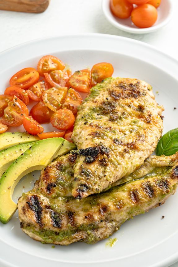 Grilled basil pesto chicken breasts served with cherry tomatoes and avocado on a white plate