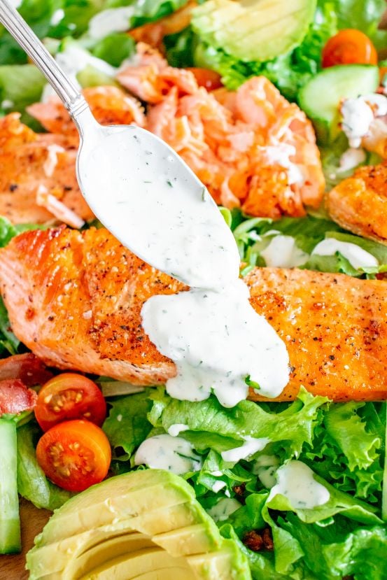 A herbed ranch dressing being spooned over a grilled salmon fillet and bacon avocado salad