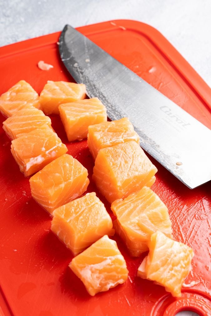 Salmon fillets being cut into even-sized chunks on a red chopping board