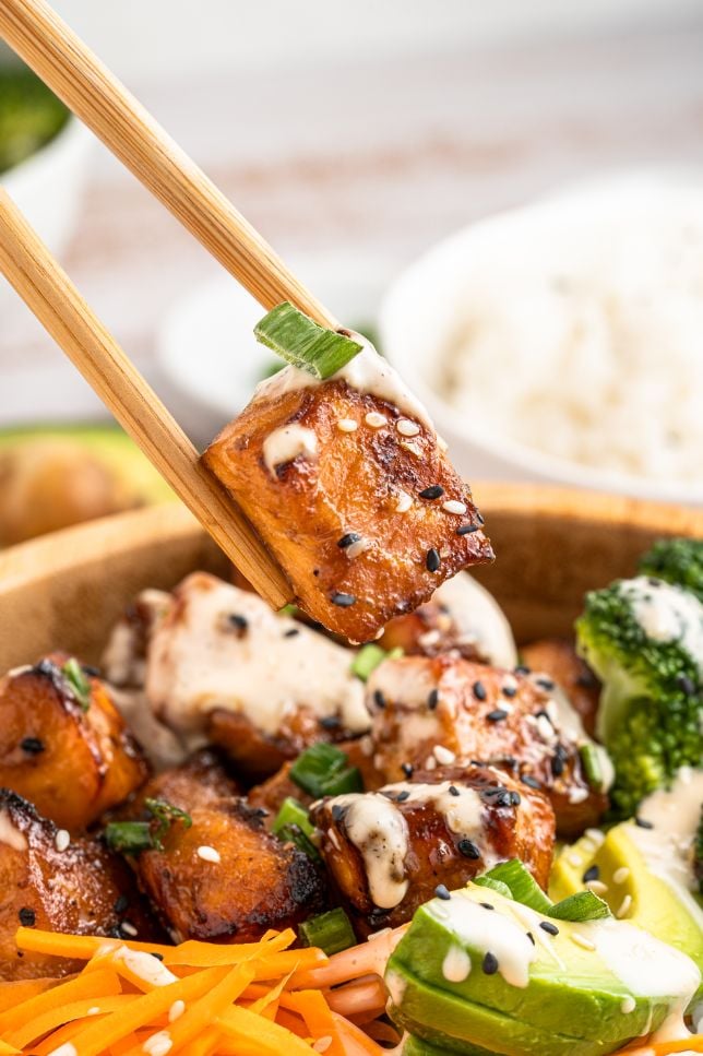 A set of chop sticks picking up a piece of cooked salmon from a rice and veggie bowl