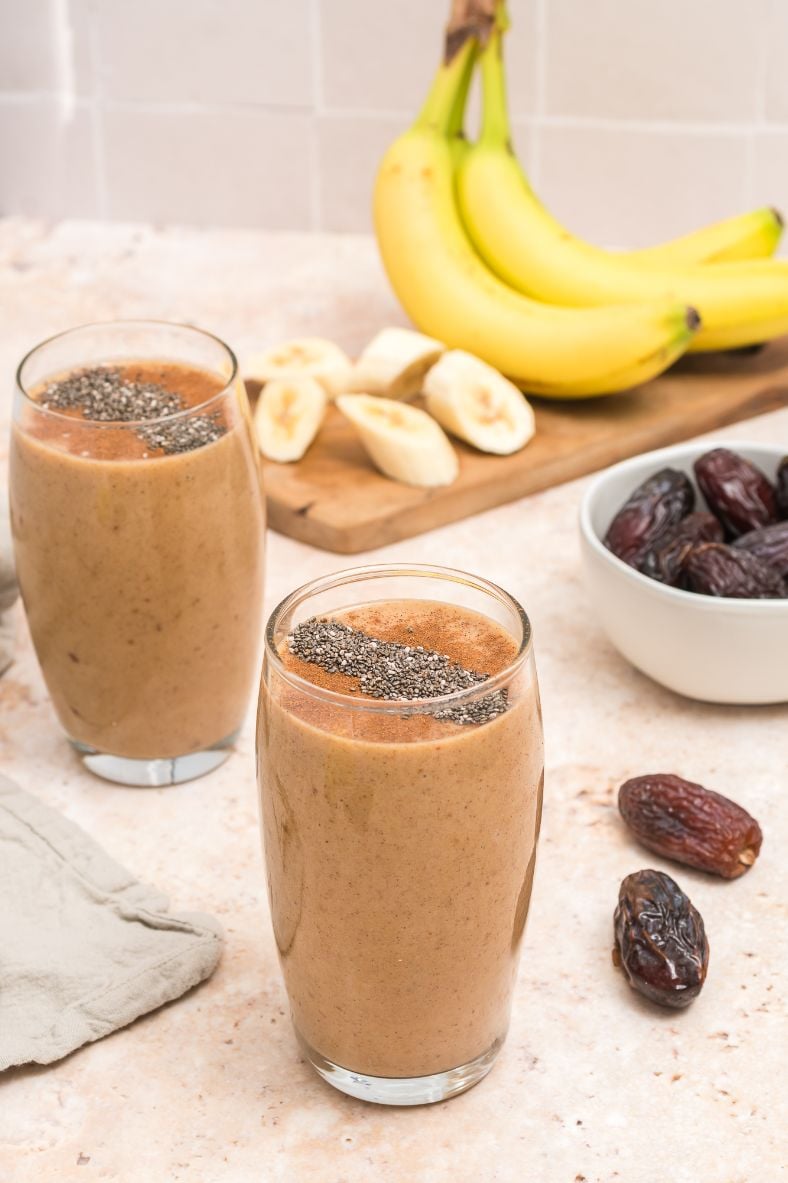 two salted caramel smoothies served in tall glasses with some bananas and dates in the background