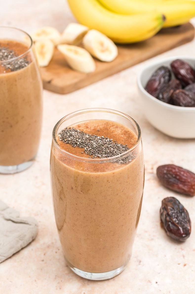 A close up shot of a caramel colored smoothie served in a tall glass with another smoothie in the background along with some fresh dates and bananas
