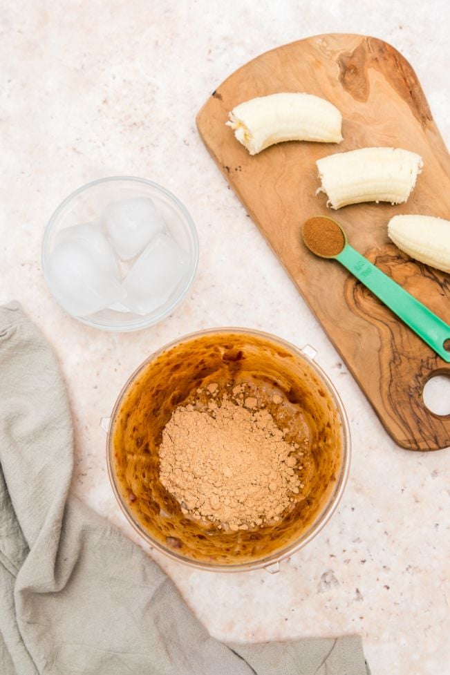 A blender jug containing blended milk and dates and extra ingredients including peanut butter powder, ice, bananas and cinnamon being added