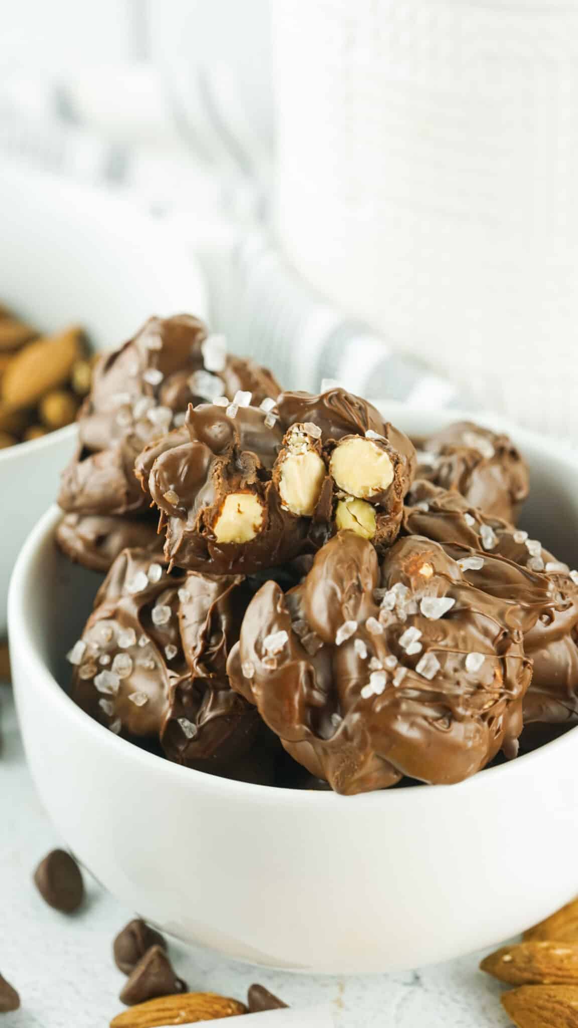 A white bowl containing a stack of chocolate covered almond clusters with one bitten through to show the almonds inside
