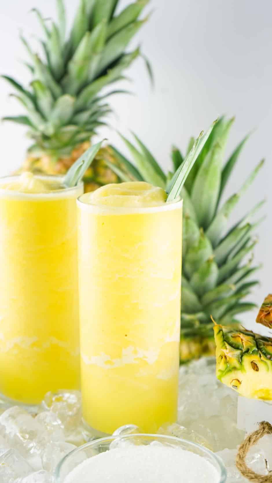 Two tall glasses filled with a frozen pineapple drink and surrounded by ice cubes and pieces of pineapple