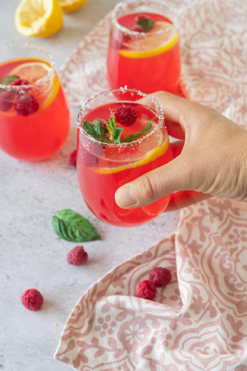 A hand reaching for a glass of a pink drink with raspberries, lemons and mint used as garnish