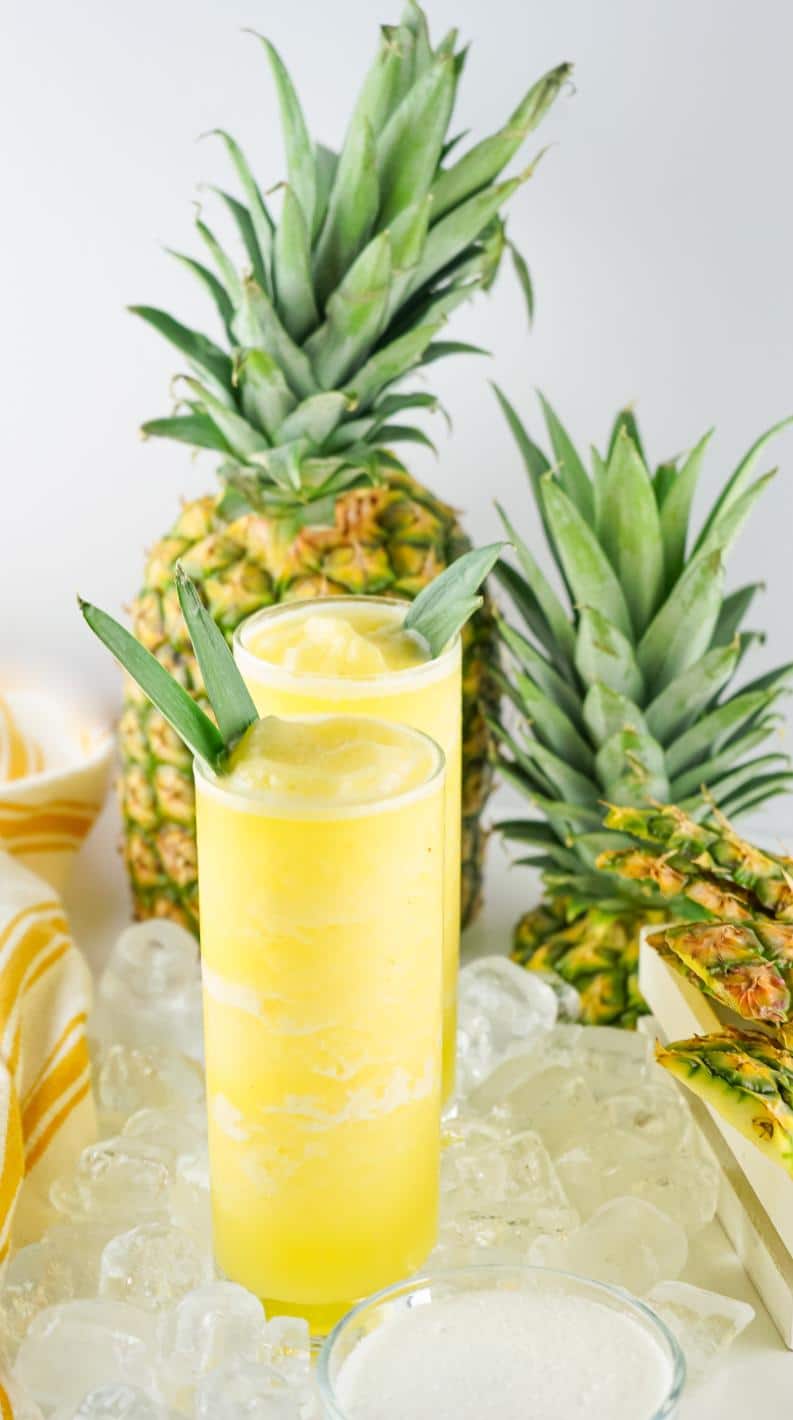 Two glasses with iced pineapple drink in them and fresh pineapples in the background