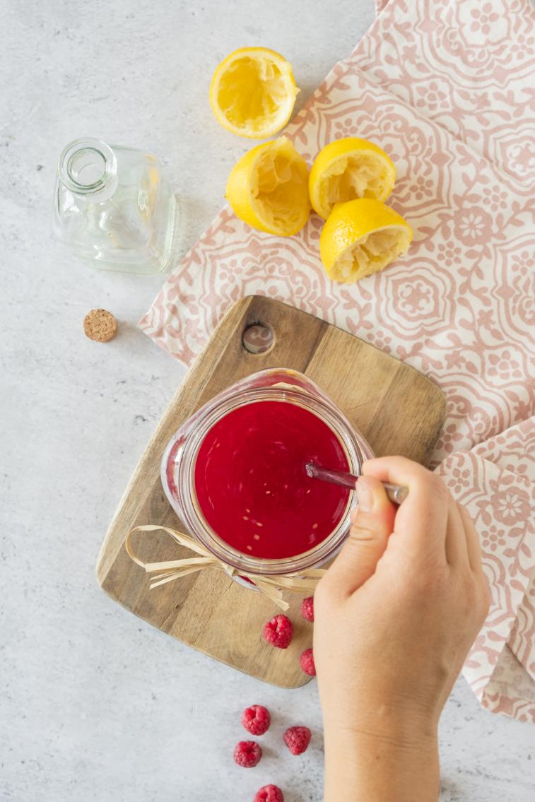 A spoon stirring a raspberry syrup in a small glass container with chopped lemons surrounding it