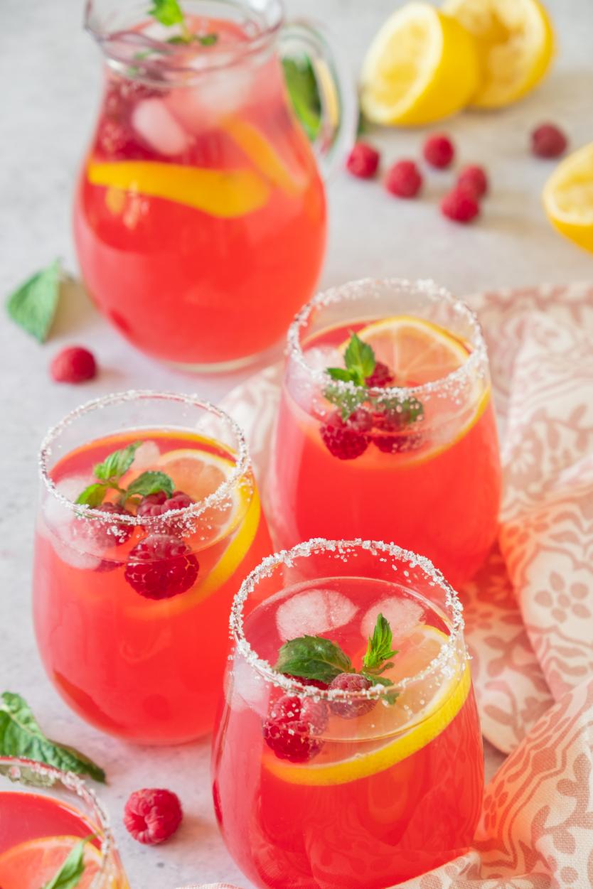 3 glasses filled with a pink drink topped with raspberries, lemon slices and fresh mint with a pitcher of the drink in the background