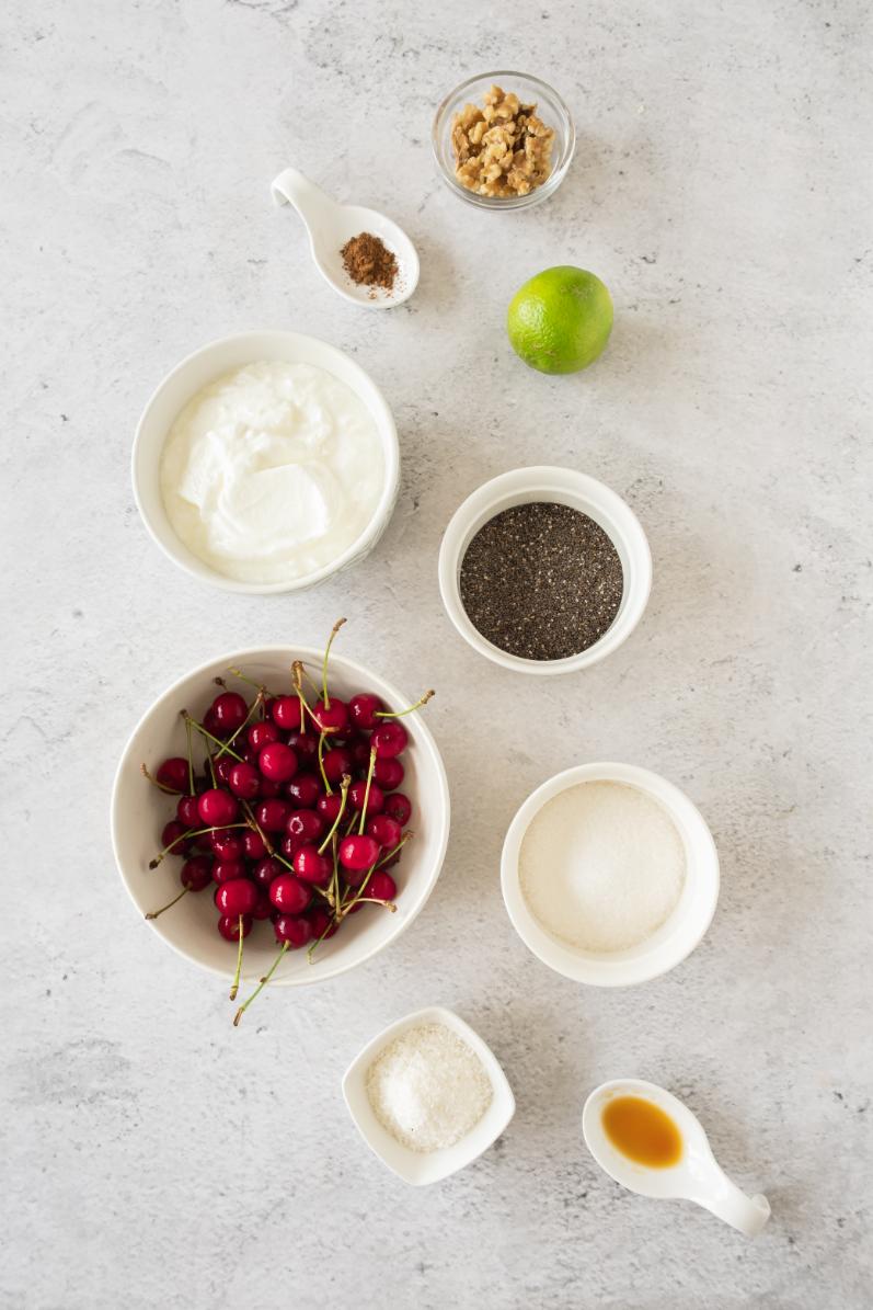 Various ingredients arranged neatly on a light surface, including cherries, yogurt, chia seeds, lime, sugar, and nuts.