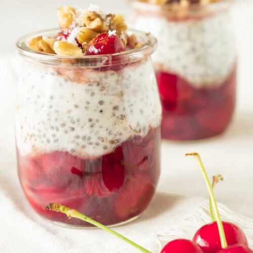 Two jars of layered cherry chia pudding with red fruit compote and topped with nuts, served on a light background with fresh cherries in the foreground.