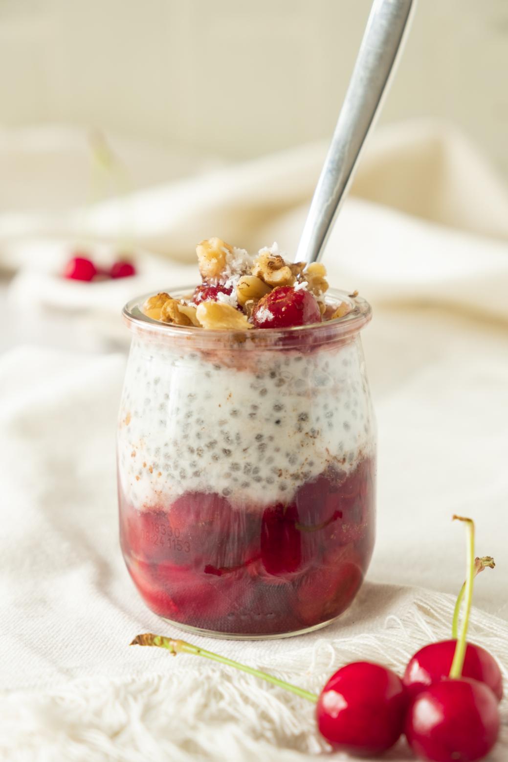 A jar of layered cherry chia pudding with a red fruit compote and a garnish of fresh cherries and chopped nuts, ready to be enjoyed as a healthy and delicious treat.