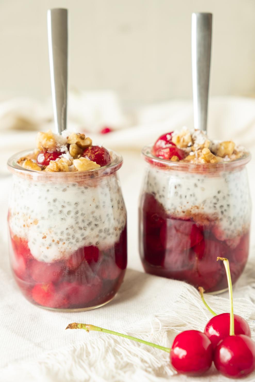 Two jars of layered cherry chia pudding, red fruit compote, and topped with crunchy walnuts, served with fresh cherries on the side.