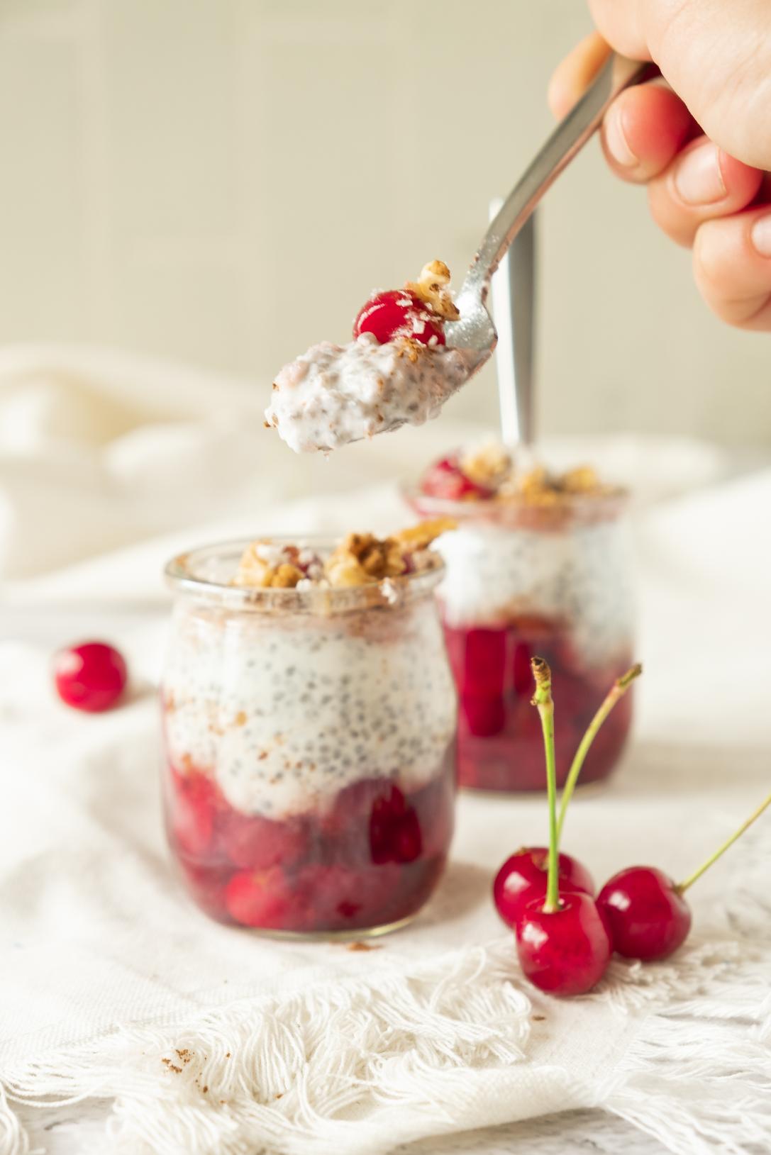 A hand holding a spoonful of creamy cherry chia pudding layered with vibrant red cherries and crunchy walnuts in a glass jar.