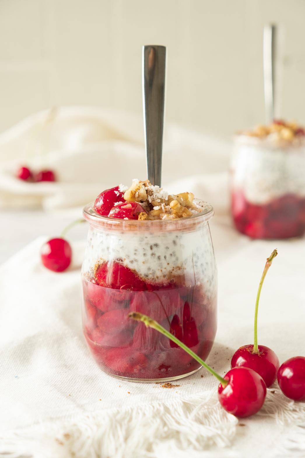 A jar of layered dessert with ruby-red cherries, creamy yogurt, and crunchy walnuts over cherry chia pudding, served with a spoon.